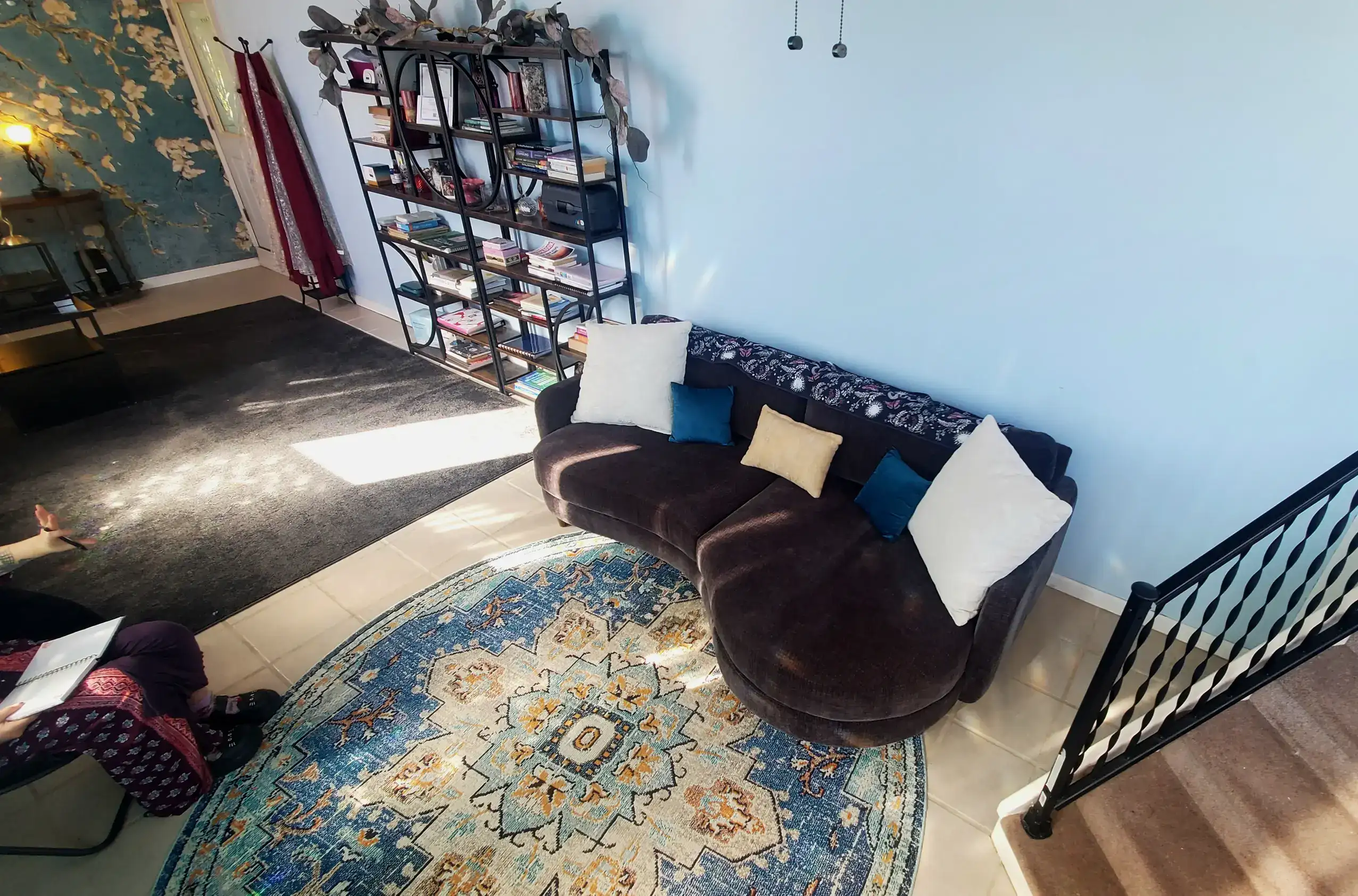 A cozy living room features a curved dark sofa with white blue and yellow pillows placed on a round patterned rug. Sunlight falls across the floor beside a tall black bookshelf filled with books while a staircase and floral wallpaper add character to the space.